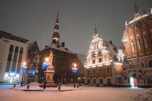 Snowy evening in Riga's Old Town with the illuminated House of the Blackheads, a statue in the square, and St. Peter's Church spire in the background. photo