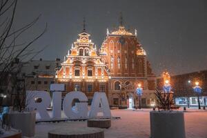 The House of the Blackheads in Riga, Latvia, glows with warm lights. Snowfall, blue lit trees, and large RIGA letters create a festive atmosphere. photo
