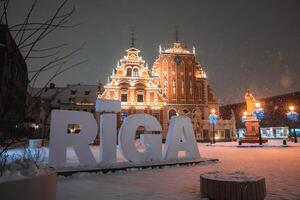 The House of the Blackheads in Riga, Latvia, glows under winter lights with snow covered ground, blue lit trees, and a statue in the square. photo