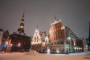 Snow covered square in Riga, Latvia, with the illuminated House of the Blackheads, St. Peter's Church spire, festive lights, and a central statue. photo