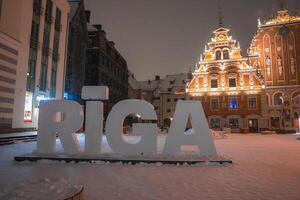 A snowy evening in Riga, Latvia, featuring the RIGA sign, the illuminated House of the Blackheads, and surrounding buildings in a serene winter setting. photo