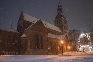Riga Cathedral in Latvia during a snowy evening. Warm streetlights highlight the red brick Gothic architecture, with snow falling and trees nearby. photo