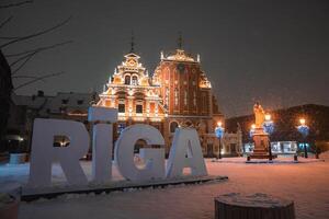 Winter evening in Riga, Latvia, with the illuminated House of the Blackheads, snow covered ground, streetlights, and a statue in a festive square. photo