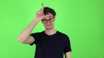 Brunette young man in t-shirt making loser gesture, L sign on forehead, teasing, accusing for defeat, expressing disrespect. Indoor studio shot isolated on green background. Concept - you lose. video