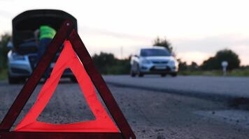 Unrecognizable sad driver in reflective vest. Male driver standing near broken car with open up hood. Red triangle to warn other road users of car breakdown or engine failure stop at countryside highway. Emergency traffic situation video