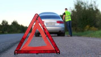 Broken silver luxury car emergency accident. Man driver installing red triangle stop sign on road. Sport automobile turned on blinkers technical problems on the road. Safety procedure when having vehicle broken down video