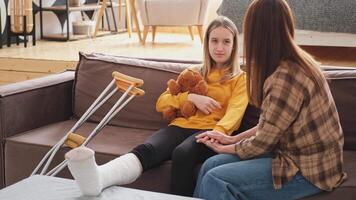 Girl with broken leg in cast sits with mother on couch at home. Sad little kid hugging teddy bear recovering from broken leg. Girl using cast crutches for quick rehabilitation. Mom supporting daughter video