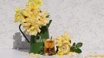 bouquet of yellow roses stands in a green vase next to a glass of tea with lemon, scattered rose petals, creating a harmonious still life on a white table, video