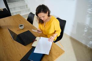 Businesswoman Working Remotely in Cozy Home Office with Laptop and Smartphone. She reviews documents and enjoys the calm workspace environment, focusing on her tasks with dedication. photo