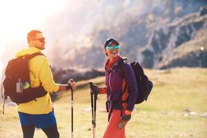 Couple hikers enjoy a scenic mountain trek in the sunshine.A man and a woman with backpacks and trekking poles walk through a green mountain landscape, smiling under the sun photo
