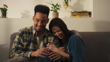 Happy international couple in casual clothes sitting and laughing on the couch hugging each other in a modern living room at home. video
