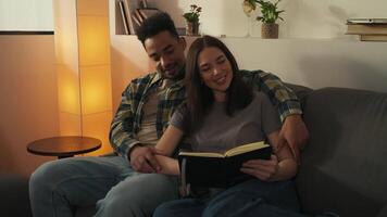 Portrait of beautiful multiracial couple sitting on the couch in a comfort atmosphere and reading book near lamp. video