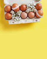 Easter eggs decorated with lace and white spring flowers in carton box on yellow background. Easter holiday, aesthetic top view of festive chicken egg in container, celebration life still photo