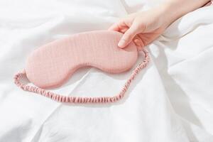Close up Pink eye mask for sleep in woman hand on white bedclothes, minimal lifestyle aesthetic flat lay. Top view sleeping mask for best sleepers, for travel, comfort relax. Rest well concept. photo