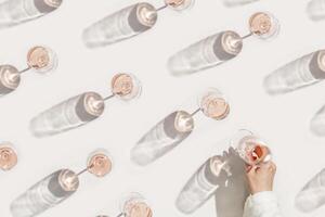 Set glasses of rose wine, home wine party Minimal concept. Top view trend pattern wih wineglass, woman hand take one, sun shadow and glare, beige pink color, alcohol drink background, flat lay photo
