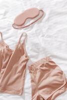 Beige female pajama and sleep eye mask on white cloth bed sheet. Top view comfort pyjamas for health sleeping. Life style photo, singlet and shorts for girl light trend color, sleepwear photo