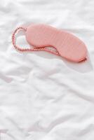 Sleeping mask pink color on bed white bedclothes background. Eye cover for best sleep. Concept of home comfort and wellbeing, dream well, comfort rest at night. Top view sleep mask, copyspace photo