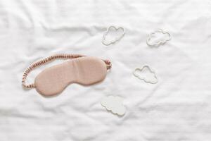 Sleeping mask beige color and clouds shapes on bed white bedclothes background. Eye cover for best sleep. Creative Concept of dream well and good night, comfort rest at night. Top view photo
