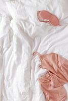 Top view pink pajama and eye sleep mask on white crumpled bedclothes, cushion and blanket. Cozy pyjamas for comfort rest at night. Flat lay from singlet, shorts, sleeping mask pastel pink color photo