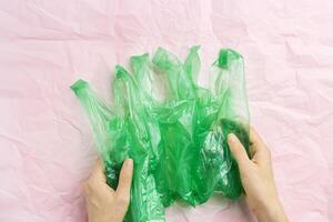 Green plastic bags in woman hands on pink crumpled paper background, conscious lifestyle concept, minimal style image. Single-use plastic ban. Eco trend to reduce disposable plastics, top photo