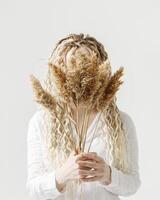 Woman holding pampas grass in front of face, light background, no face trend concept. Female with long curly hair dreadlocks hiding behind bouquet dry flower. Creative autumn visual. Fall concept photo