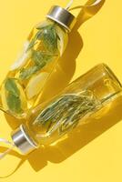 Top view closeup two glass bottles, water drink detox with lemon and mint leaves, and rosemary, at sunlight on yellow color background. Infused water, wellness trend. Refreshing summer drink photo