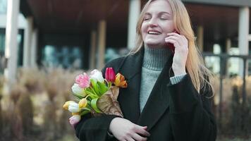 Romantic woman joyful blogger having fun holding flowers using a smartphone shooting self portrait on front camera call enjoying event holding colorful tulips gift case in package video