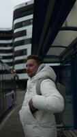 Young stylish man with smart phone on bus stop and typing sms stand at public transport bus station taking bus with headphones and mobile smart phone in winter or autumn day with backpack video