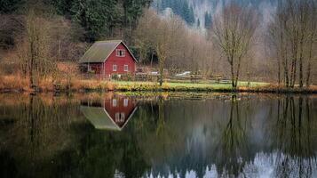A tranquil scene features a rustic red cabin nestled among trees, reflected beautifully in calm water. This picturesque landscape offers serenity and natural beauty. photo
