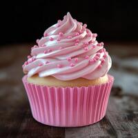 A pink cupcake with pink frosting and pink sprinkles on top. The cupcake is sitting on a wooden table photo