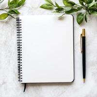 Open blank notebook with pen on marble surface, bordered by green leaves, perfect for writing or journaling concepts. photo