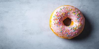 A delightful pink frosted donut adorned with colorful sprinkles sits atop a textured grey background, perfect for showcasing sweetness and indulgence in culinary art. photo
