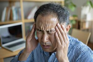 Stressed Asian man experiencing a headache at home office, showcasing a common health issue, feeling discomfort and needing relief The image captures the tension and pressure associated with stress photo