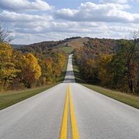 A picturesque country road stretches into the distance, flanked by vibrant autumn trees ablaze with color The yellow lines guide the eye towards a gentle, rolling hill in the horizon, topped with a photo