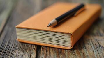 A simple composition, capturing the essence of study and reflection, features an orange notebook resting upon a weathered wooden surface A sleek, dark pen lies gracefully atop the notebook, hinting photo