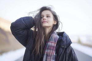 A serene portrait captures a young woman against a bright, slightly blurred backdrop Her long, wavy brown hair flows loosely, framing her face as she gazes upwards with a gentle smile She is dressed photo
