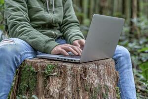 Amidst verdant foliage, a person finds their workspace, a silver laptop perched atop a weathered tree stump Sunlight filters through the canopy, dappling the scene in a soft, ethereal glow Fingers photo