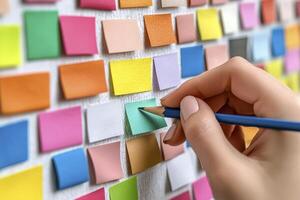In a close-up image, a hand delicately holds a sharpened pencil, poised above a vibrant array of colorful sticky notes meticulously arranged on a textured white surface Each note represents a photo