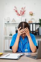 A healthcare worker in blue scrubs appears stressed while sitting at a desk with medical documents and equipment in an office. photo