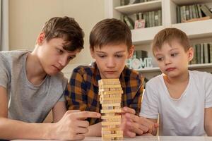 Three boy brothers are playing a board game made of wooden rectangular blocks, pulling pieces out of the tower. photo