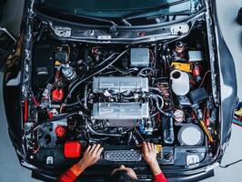 A person working on a car engine, focusing on mechanical components and tools under the hood in a well-lit garage setting. photo