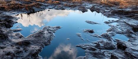 A shallow puddle reflecting clouds, surrounded by muddy terrain and dry grass, showcases a serene natural landscape. photo