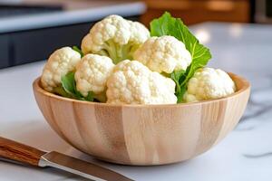 Fresh cauliflower florets in wooden bowl on kitchen counter with knife photo