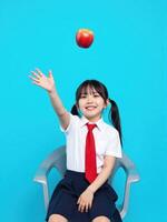 A little girl in a school uniform is sitting on a chair and holding an apple photo