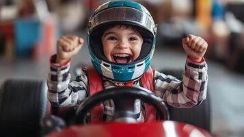 A young boy is racing with a car on a track He wears a helmet and looks happy He is cheering with his hands raised in the air photo