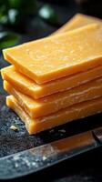 Bright yellow cheese stacked on a dark surface with a knife nearby in a kitchen setting photo