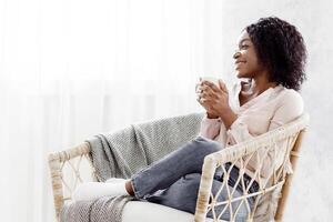 Perfect Day Off. Cheerful Black Woman Relaxing In Wicker Chair With Coffee, Looking Aside, Sode View With Free Space, Panorama photo