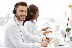 Handsome call center operator smiling at camera and his coworkers talking to clients at office, copy space photo