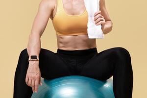 Equipment for training and coach online. Adult smiling muscular slim lady in sports uniform holds towel and sits on fitness ball, isolated on orange background, panorama, free space, studio shot photo