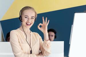 Beautiful female helpline consultant looking at camera and showing OKAY gesture in modern office. Call centre operator expressing positivity and her collagues at open workspace photo